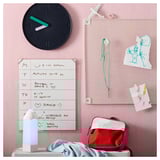 Organized kids area: wall clock, whiteboard, bulletin board with drawings, red backpack, folded blanket, blue lamp.