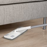 White mop with grey microfiber pad leans against modern grey upholstered furniture on wooden floor.