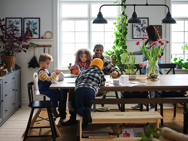 Happy family crafting together at a cozy wooden table with bench, surrounded by plants and large windows.