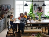 Happy family crafting together at a cozy wooden table with bench, surrounded by plants and large windows.