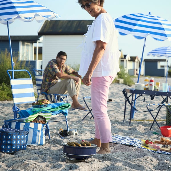 Elderly woman grilling on beach, pink pants, sausage on small grill.