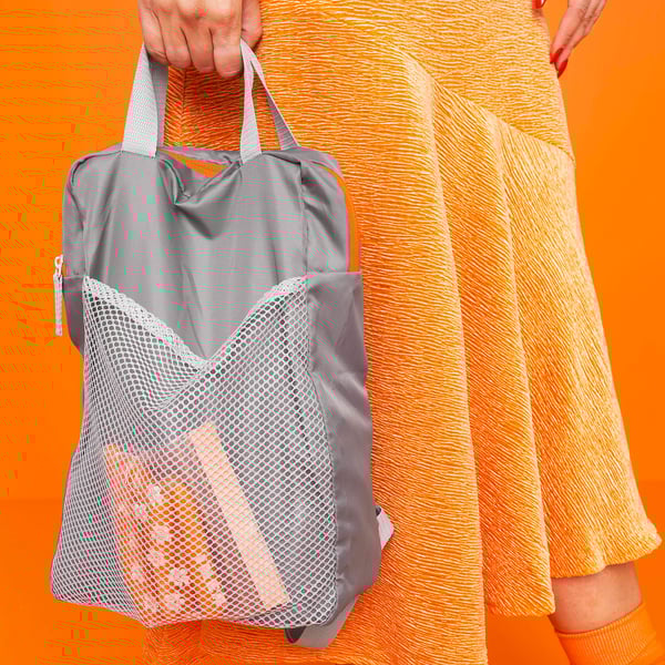 Person holding gray PIVRING backpack with mesh pocket. Orange, white outfit, bright orange background.
