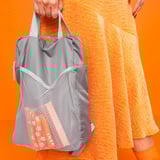 Person holding gray PIVRING backpack with mesh pocket. Orange, white outfit, bright orange background.