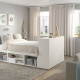 Minimalist bedroom with PLATSA bed, sheer curtains, neutral bedding, storage boxes, books, chair, wall art, and rug.