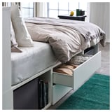 A white PLATSA bed with storage drawers, neatly made with grey and white bedding. Green rug beneath.