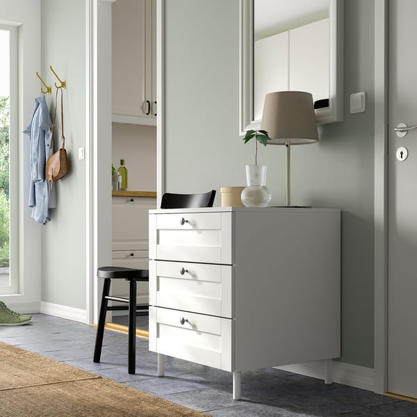 Modern bedroom with white PLATSA dresser, grey cabinet, lamp, black stool, and golden hooks for clothes.