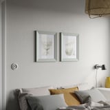 Two grey PLOMMONTRÄD frames with floral art hang above a bed with white & mustard pillows, creating a cozy atmosphere.