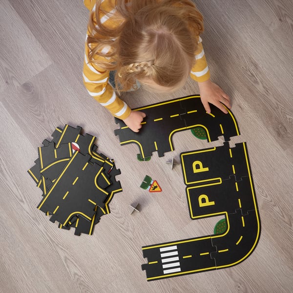 Little girl playing with RÄVUNGE car track puzzle on the floor, assembling road pieces and traffic signs, enhancing problem-solving and creativity.