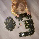 Little girl playing with RÄVUNGE car track puzzle on the floor, assembling road pieces and traffic signs, enhancing problem-solving and creativity.