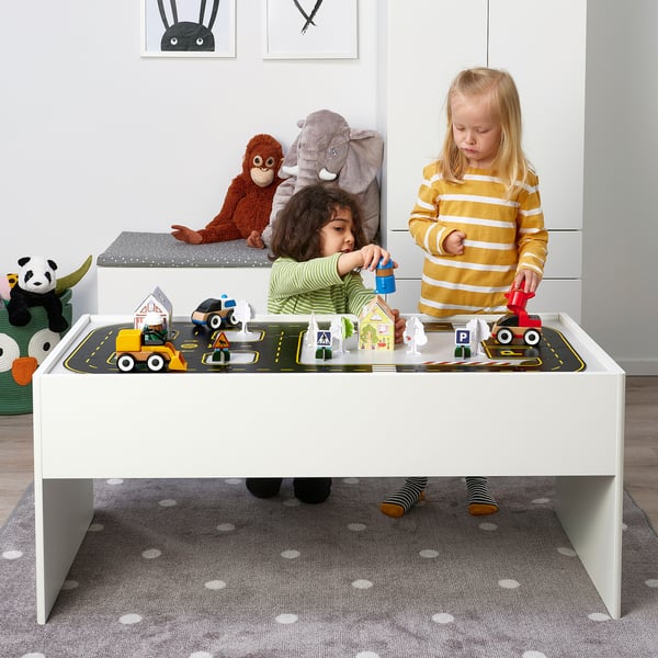 Kids play with toy car track set on table, including construction vehicles and signs, promoting creativity and motor skills.