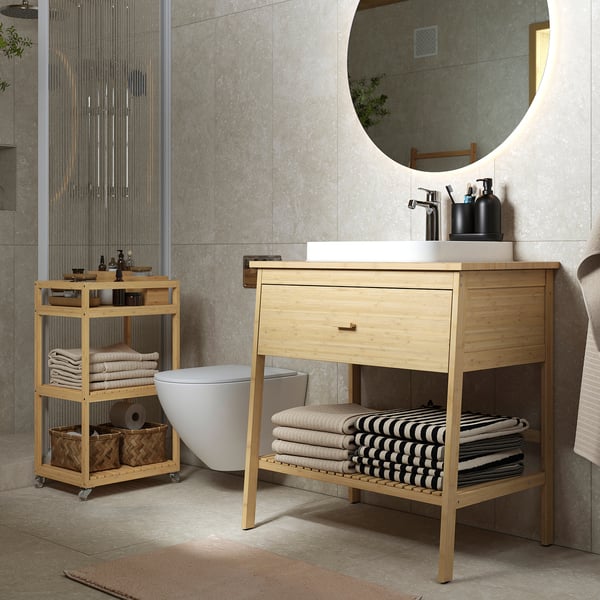 Modern bathroom with light wooden sink vanity, white basin, round mirror, and mobile storage cart with towels and toiletries.