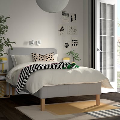 Grey RÅHOLMAR bed with 5 comfort zones, black/white pillows, striped throw, wall art, round light, wooden bedside table, plant, window, rugs.
