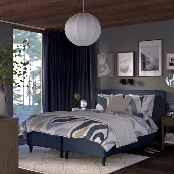 Modern grey RÅHOLMAR bed with wooden accents, abstract linens, pendant lamp, wall frames, blue curtains, and a plant.
