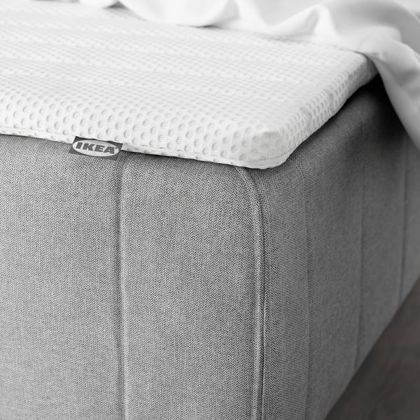 A white mattress with a textured topper, visibly supported by a grey frame, offering comfort and spine alignment in a bedroom setting.