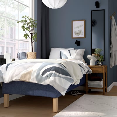 Modern bedroom with light blue RÅHOLMAR bed, white/gray bedding, wooden bedside table, lamp, plant, dark blue walls, mirror, robe.