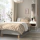 Modern bedroom with beige RÅHOLMAR bed, patterned bedding, white side table, mirror, decorative plants, and large window letting sunlight in.