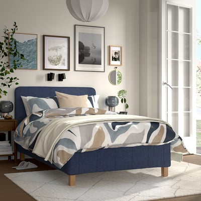 Modern bedroom with RÅHOLMAR bed, dark blue, patterned comforter, pillows, art, rug, plant.