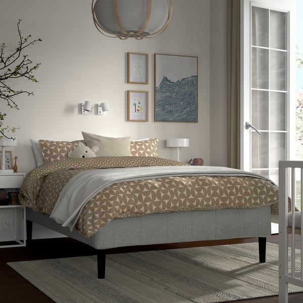 Modern bedroom with RÅHOLMAR bed, beige-white bedding, teddy bear, lamp, art, side table with books, large window.