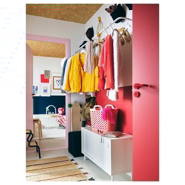 Bright pink room with yellow and red clothing on hangers, white storage unit, pink laundry baskets, and a jute and wool rug on the floor.