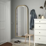 Gold RÅMEBO mirror against gray wall, reflecting door, dresser with blue jacket, small stool.