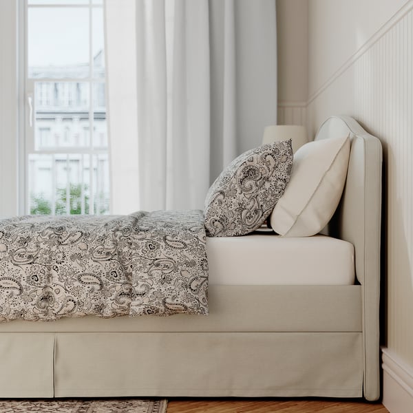 White bed with patterned cover and pillows, large window with view to buildings, soft lighting, beige bed skirt.