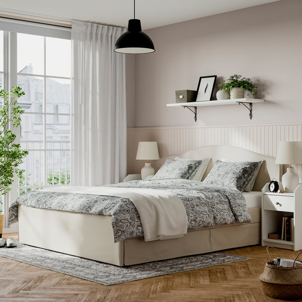 RAMNEFJÄLL bed in beige with pattern, small bedside table, windows with sheer curtains, plant, & wall shelf.