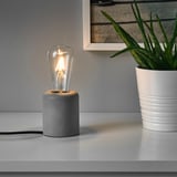 A small table lamp with a concrete base sits on a white surface, its glowing bulb providing warm, soft light. Beside it, a potted plant adds greenery.