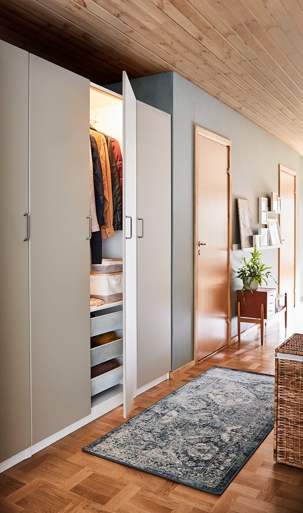 Modern closet with REINSVOLL white door revealing organized clothes and storage. Wooden floor, light blue rug, plant, and bookshelf in background.