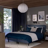 Modern bedroom with large bed, dark bedding, white pillows, lamp, dark curtains, art, wood floors, side tables, green plant.