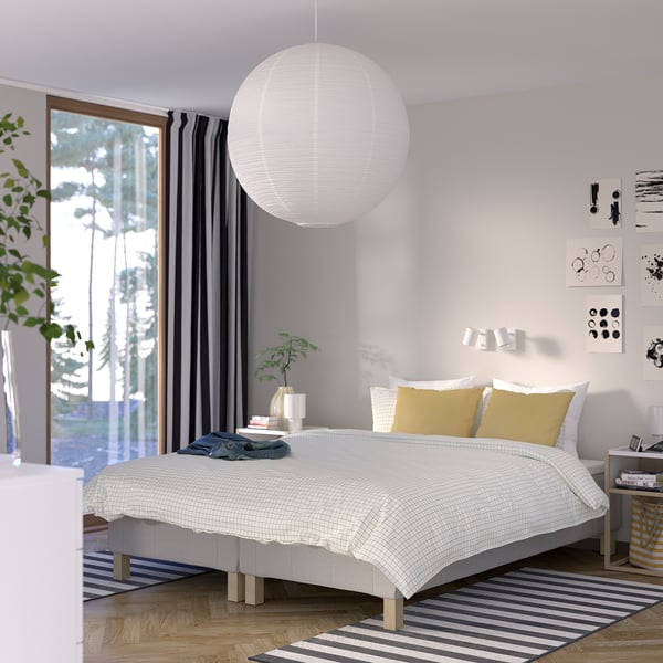 Modern minimalist bedroom with large white bed, mustard pillows, striped rug, pendant lamp, side table with books, and a large window with curtains.