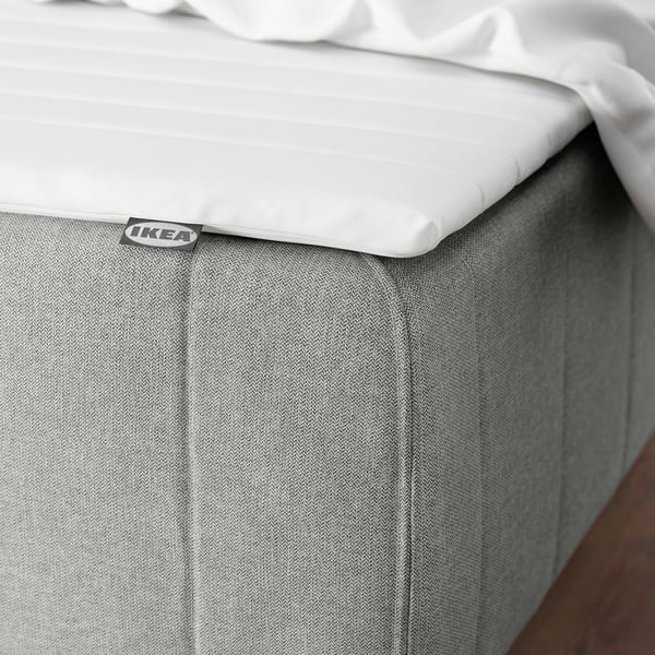 A neatly made bed with a white mattress, grey fabric bed skirt, and a visible label.