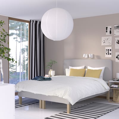 Modern minimalist bedroom with a large bed. White bedding, yellow pillows. Large window, grey curtains. Decorative plants and art. Hanging white bulb.
