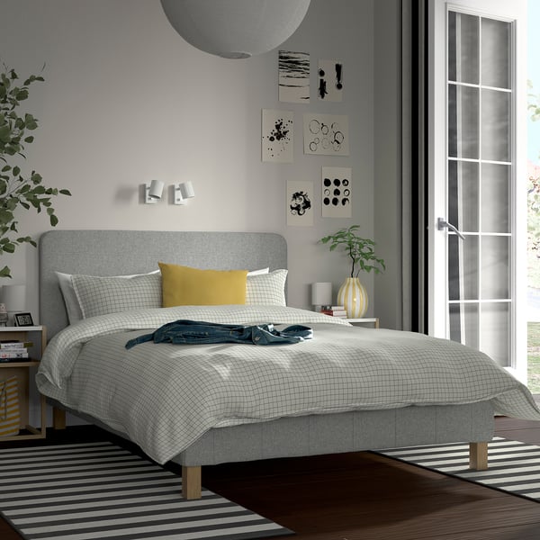 Modern bedroom with grey RENFJÄLLET bed, yellow pillow, side tables, plants, lamps, abstract art, striped rug, large window.