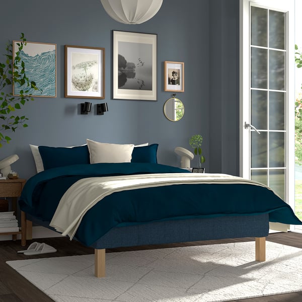 Modern bedroom with RENFJÄLLET bed in dark blue, wooden legs, art-adorned wall, rug, lamp, books, plant.