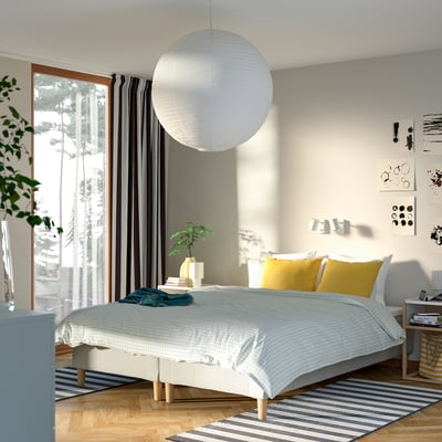 Modern bedroom with large window, grey curtains, white bed with yellow pillows, striped rug, round light fixture, small plant, and wall art.