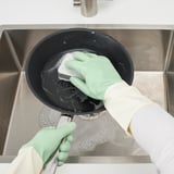 A person wearing green dish gloves washes a black pan in a stainless steel sink, demonstrating kitchen cleaning.