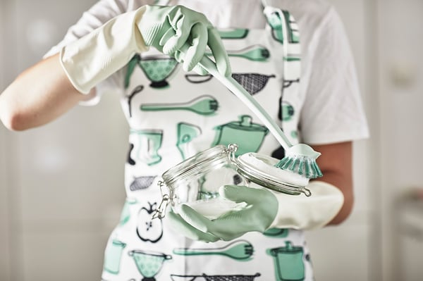 Person wearing green and white dish gloves, apron with kitchen designs, holding a glass jar with a brush, cleaning.