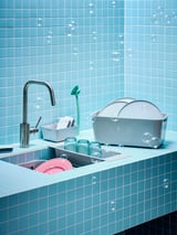 Blue sink with silver faucet, green cleaning tools, pink/blue dishes, blue tiled wall.