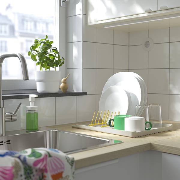 Bright kitchen sink area with plates drying in colorful holder, green soap dispenser, plant, and floral cloth draped nearby.