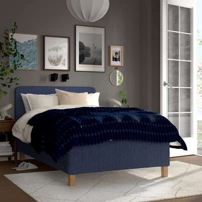 Blue bed with checkered blanket, 2 white, 1 beige pillow, grey wall art, french door, small rug, dark floor.