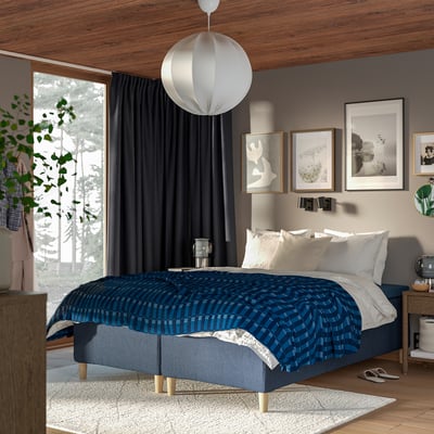 Bedroom with RISHÖJDEN bed, blue spread, dark curtains, wood ceiling/floor, hanging lamp, window, wall art, wooden nightstands.