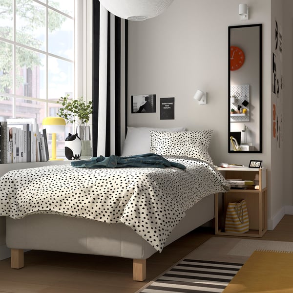 Modern bedroom with RISHÖJDEN bed, dotted bedspread, gray pillows. Yellow lamp, books, plant, mirror, wooden table, striped bag. Minimalist decor.
