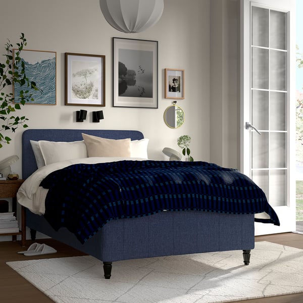 A modern blue bedroom with RISHÖJDEN bed, dark patterned spread, pillows, framed wall art, mirror, large window, and rug.