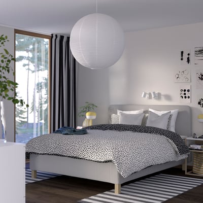 Modern bedroom with RISHÖJDEN bed, grey polka-dot comforter, white pendant light, striped rug, plants, and large windows with grey curtains.