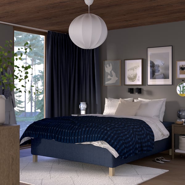 Cozy bedroom with RISHÖJDEN bed, dark blue cover, wooden nightstands, hanging lamp, large window, plants, and wall art.