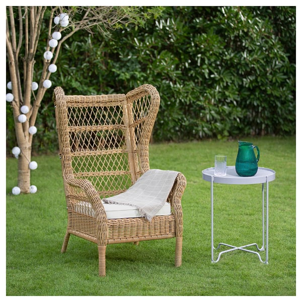 Beige wicker RISHOLMEN chair with cushion on lawn, side table with green pitcher and glass, greenery, lights.