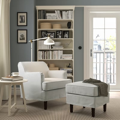 Cozy reading nook: grey chair, striped cushion & ottoman, beside bookcase & lamp, under window.