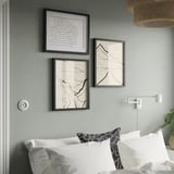 Three black art frames above bed: abstract, winding, steep, wavy. White pillows, patterned pillow, wall light, basket, plant.