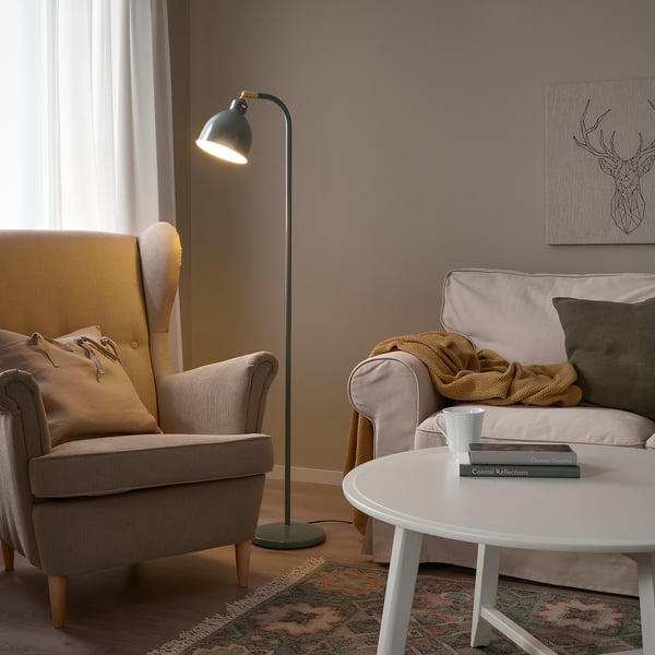 Cozy living room with RÖDFLIK lamp, beige chair, sofa, throw blanket, books, and rug. Lamp stands tall near window, casting warm light.
