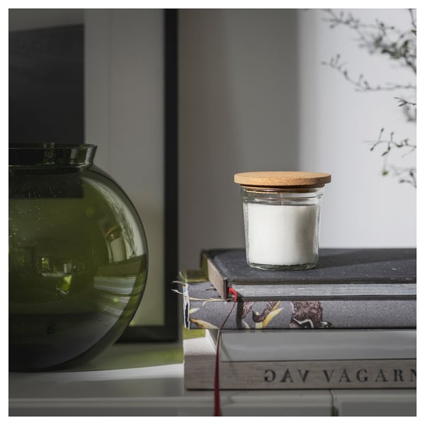 Small round glass jar with white candle and cork lid sits atop books next to green vase.